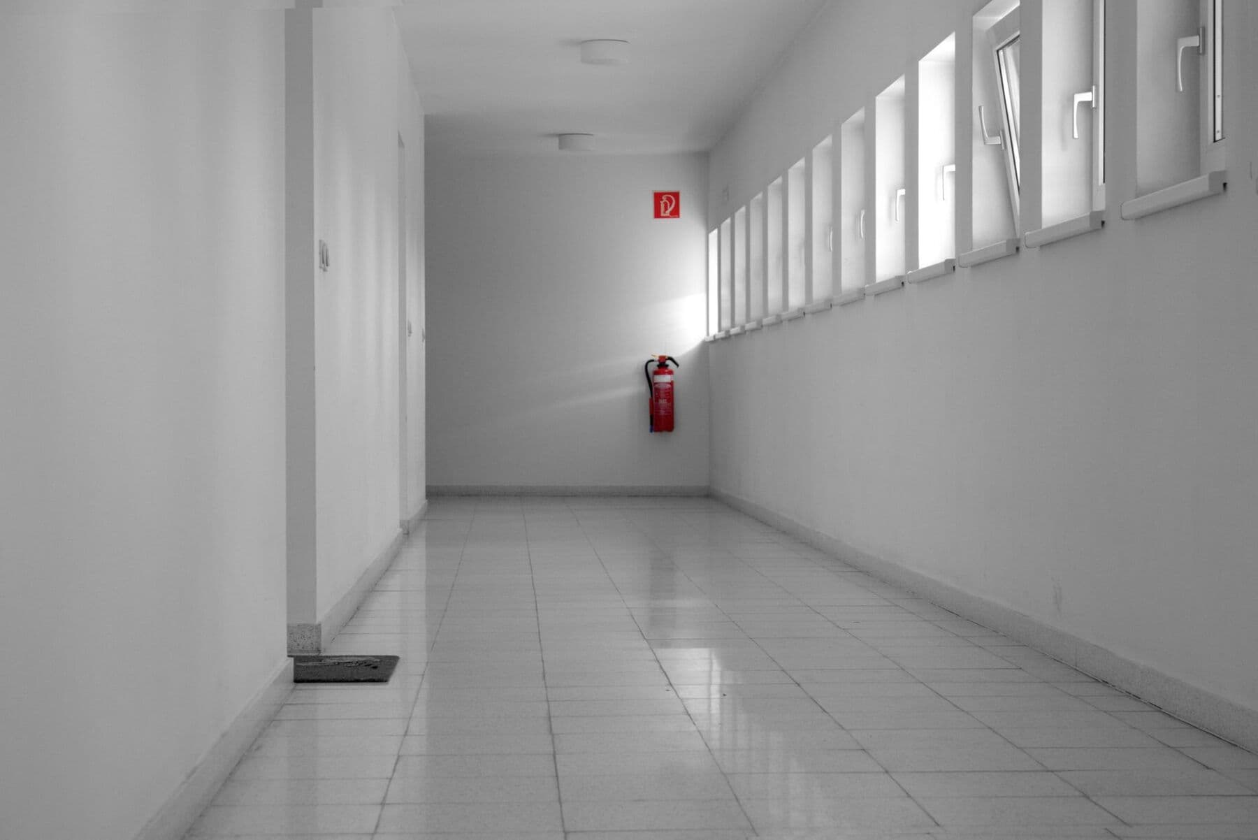 A long, empty white corridor with a row of small windows on one side and a red fire extinguisher at the far end, in black and white.